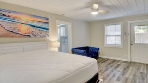 Une chambre avec un grand lit, un ventilateur de plafond, un tableau représentant une plage, une chaise bleue et une fenêtre avec des stores.