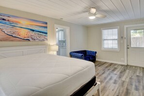 Una camera da letto con un letto grande, una ventola a soffitto, un quadro raffigurante una spiaggia, una sedia blu e una finestra con persiane.