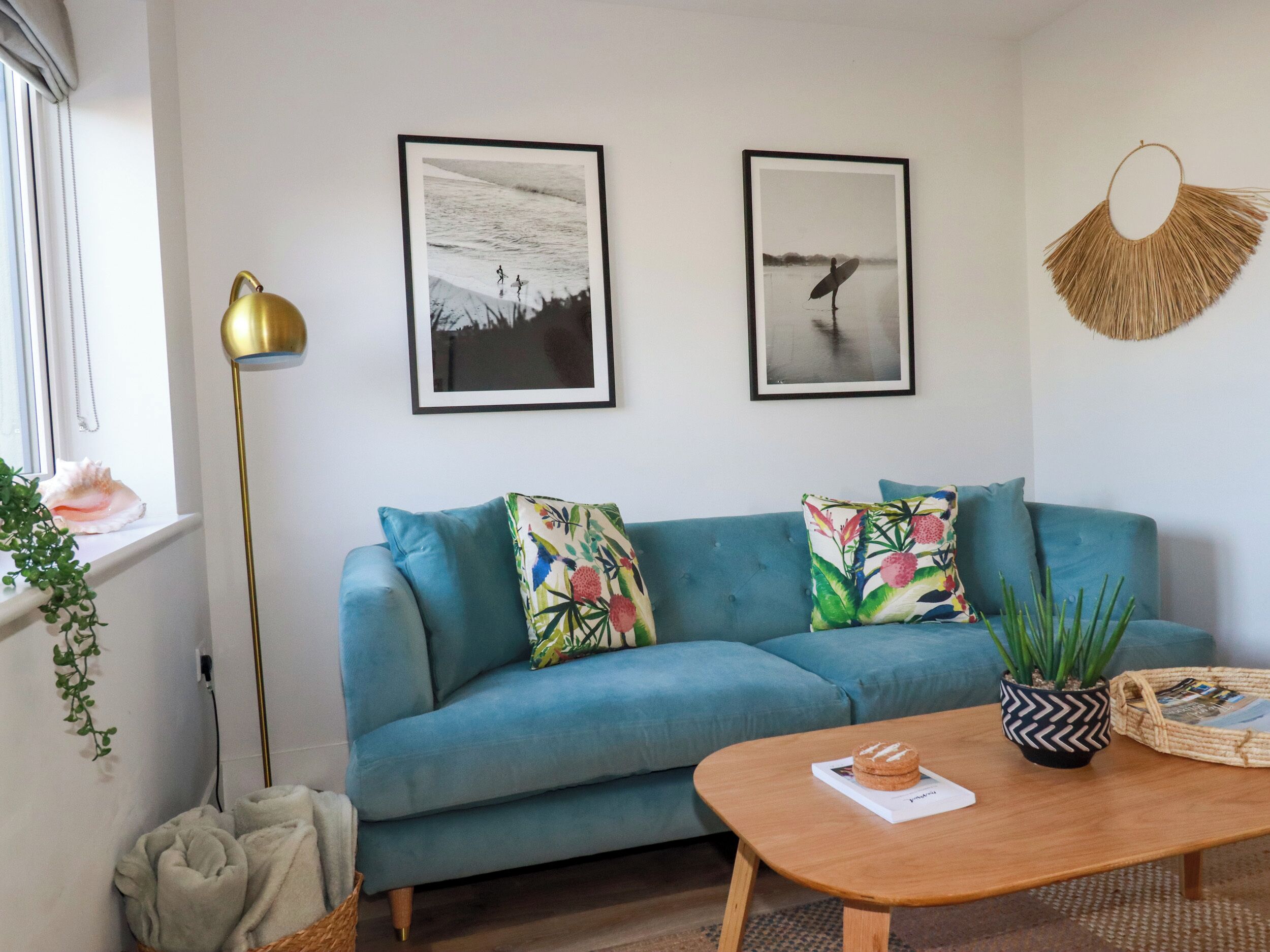 A living room with a blue sofa, two framed pictures, a floor lamp, a coffee table with a book, and a woven wall hanging.