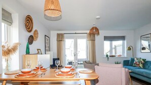 Ein modernes Wohnzimmer mit einem Holzesstisch, gedeckt für vier Personen, einem blauen Sofa und einem großen Fenster, das natürliches Licht hereinlässt.