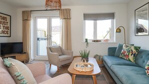 Ein Wohnzimmer mit Sofa, Sessel und Couchtisch. Ein Fernseher steht auf einem Schrank und ein Fenster ist mit Vorhängen drapiert.