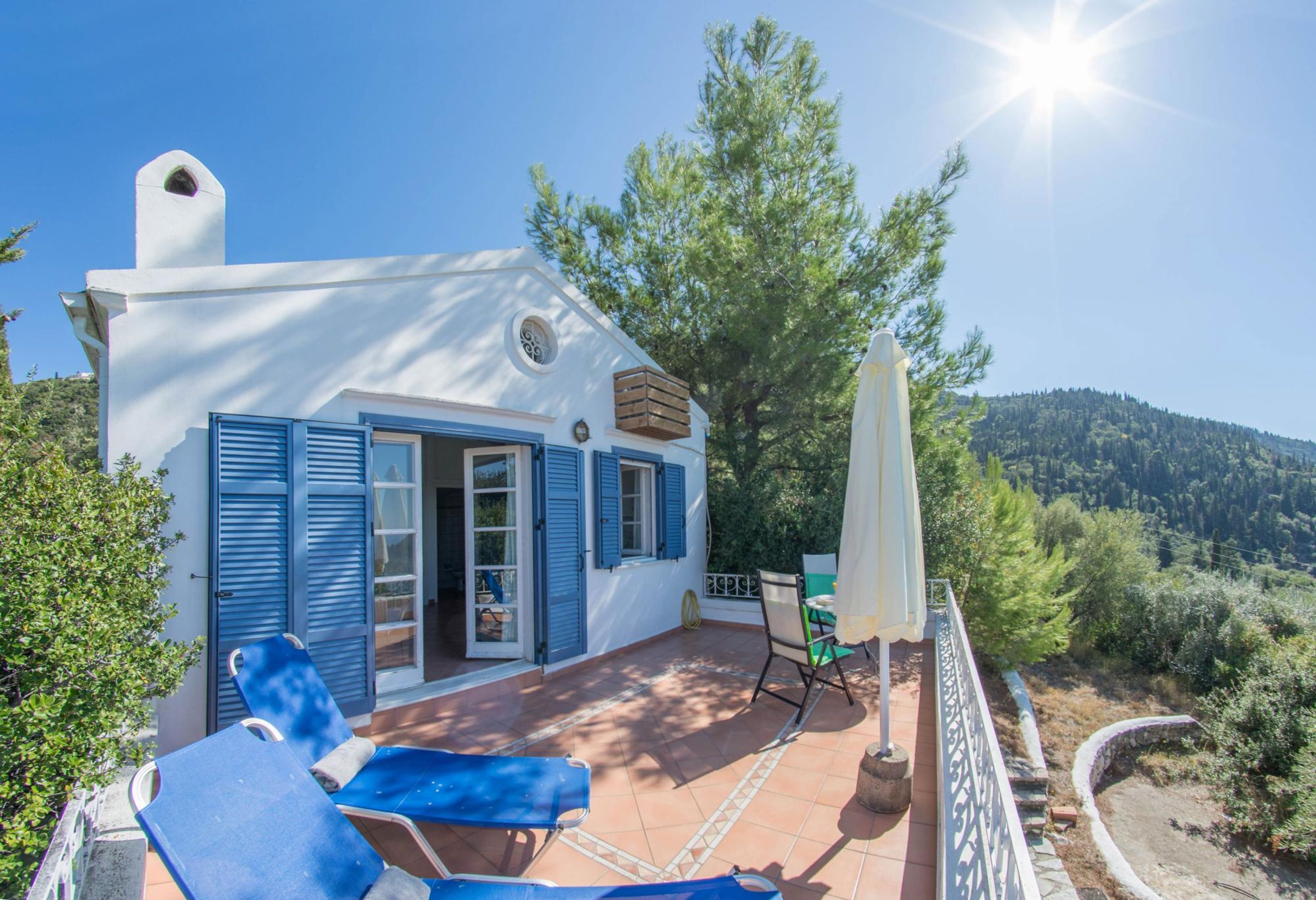 A white building with blue shutters, a patio with a table and chairs, and a view of a forested hillside.