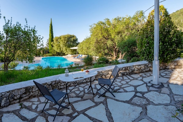 A stone patio with a table and chairs, a swimming pool, and lush greenery.