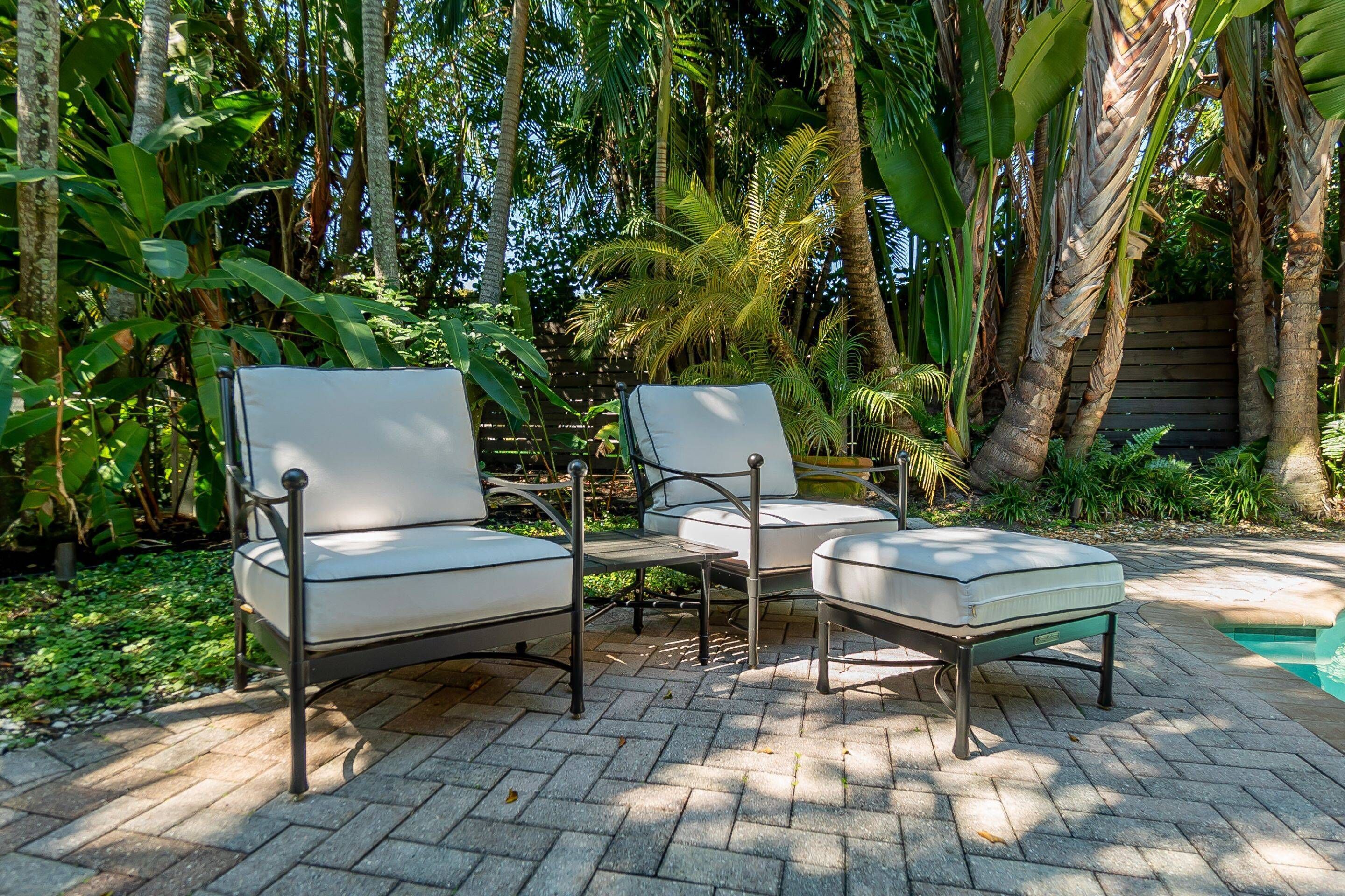 Een terras met twee witte fauteuils en een kleine tafel, omringd door weelderige tropische planten en een houten hek.