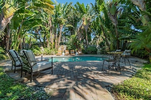 Area piscina con sedie a sdraio e un tavolo, immersa in una rigogliosa vegetazione tropicale sotto un cielo azzurro.