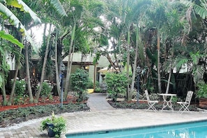 A poolside area with a pathway leading to a covered seating area surrounded by lush tropical vegetation.