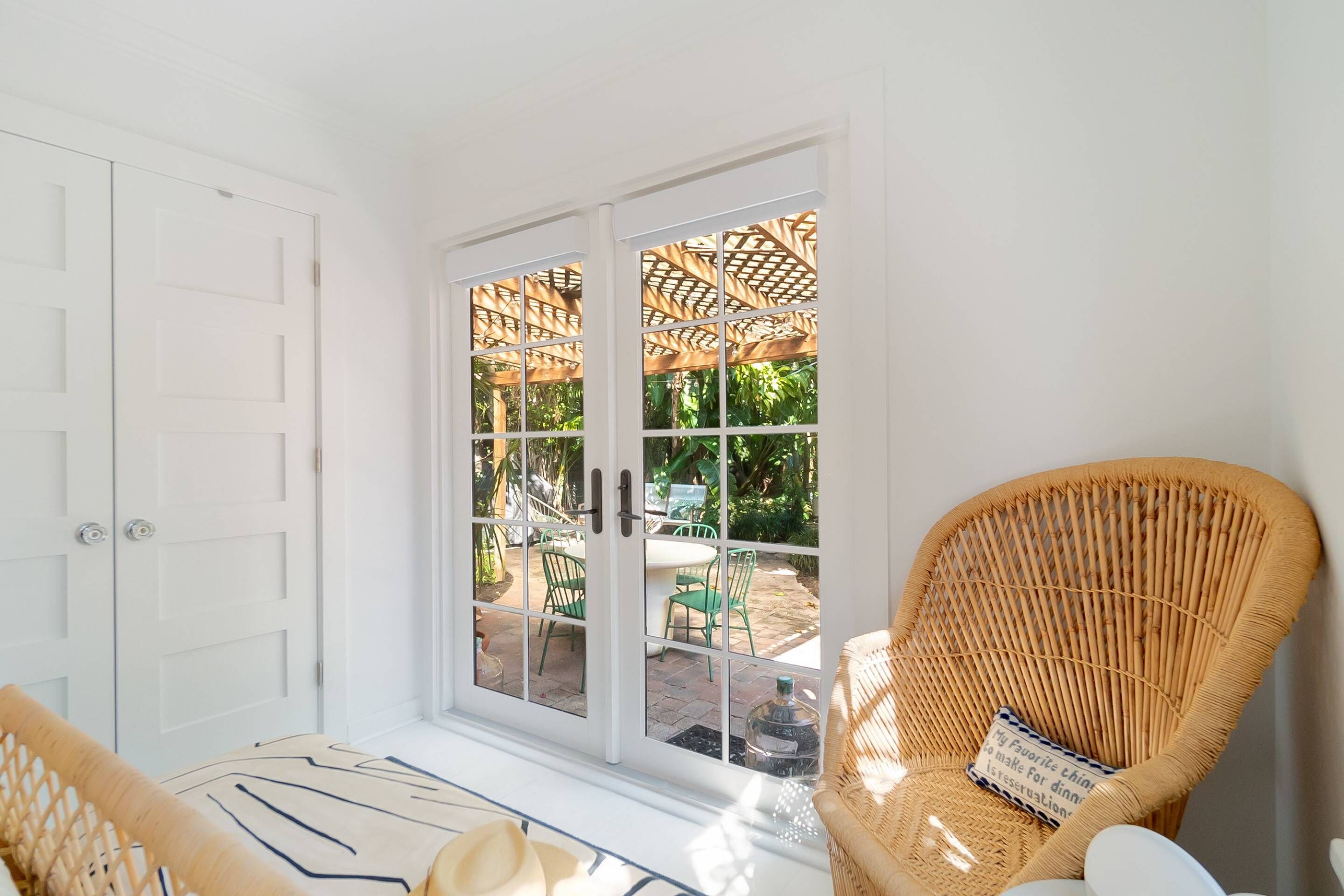 A room with a white door, a wicker chair with a cushion, and a view of an outdoor patio with a table and chairs.