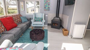 A cozy living room with a grey sofa, a light blue armchair, a patterned ottoman, a black wood-burning stove, and a white radiator.
