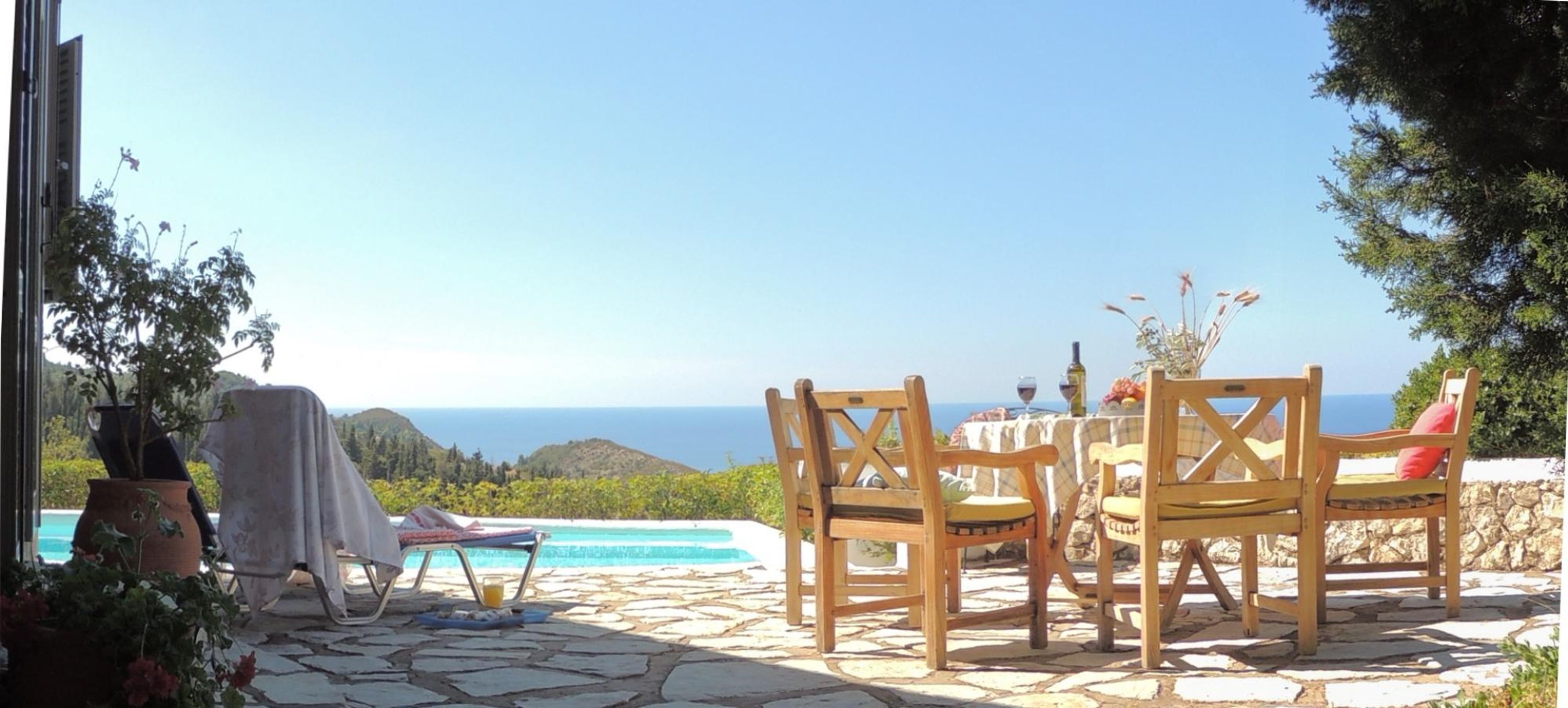 A patio with stone flooring, wooden chairs, a table, and a view of the sea.