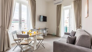 Una sala de estar moderna con un sofá, una mesa de comedor con sillas, un televisor de pantalla plana montado en la pared y ventanas grandes con cortinas.