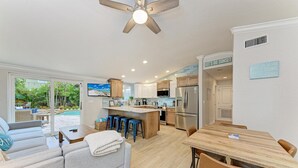 A modern kitchen with a central island, a dining area with a wooden table and chairs, and a living area with a sofa and a ceiling fan. - Beach Bliss managed by Beach Retreats (Holmes Beach)