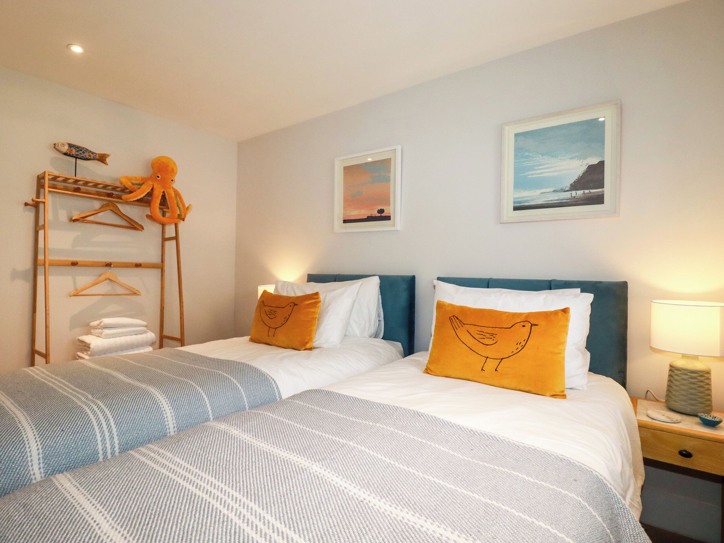 A hotel room with two beds, each with a blue headboard and white bedding. There are two pillows with bird designs, a wooden rack with towels, and two framed pictures on the wall.