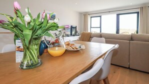 A modern living room with a wooden dining table, white chairs, a beige sofa, and a large window with curtains. - 11 Ebbtide (Newquay)