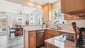 A kitchen with wooden cabinets, a granite countertop, a dishwasher, and a dining area with a table and chairs. - Beach Rays by Ryson Vacation Rentals (Galveston)