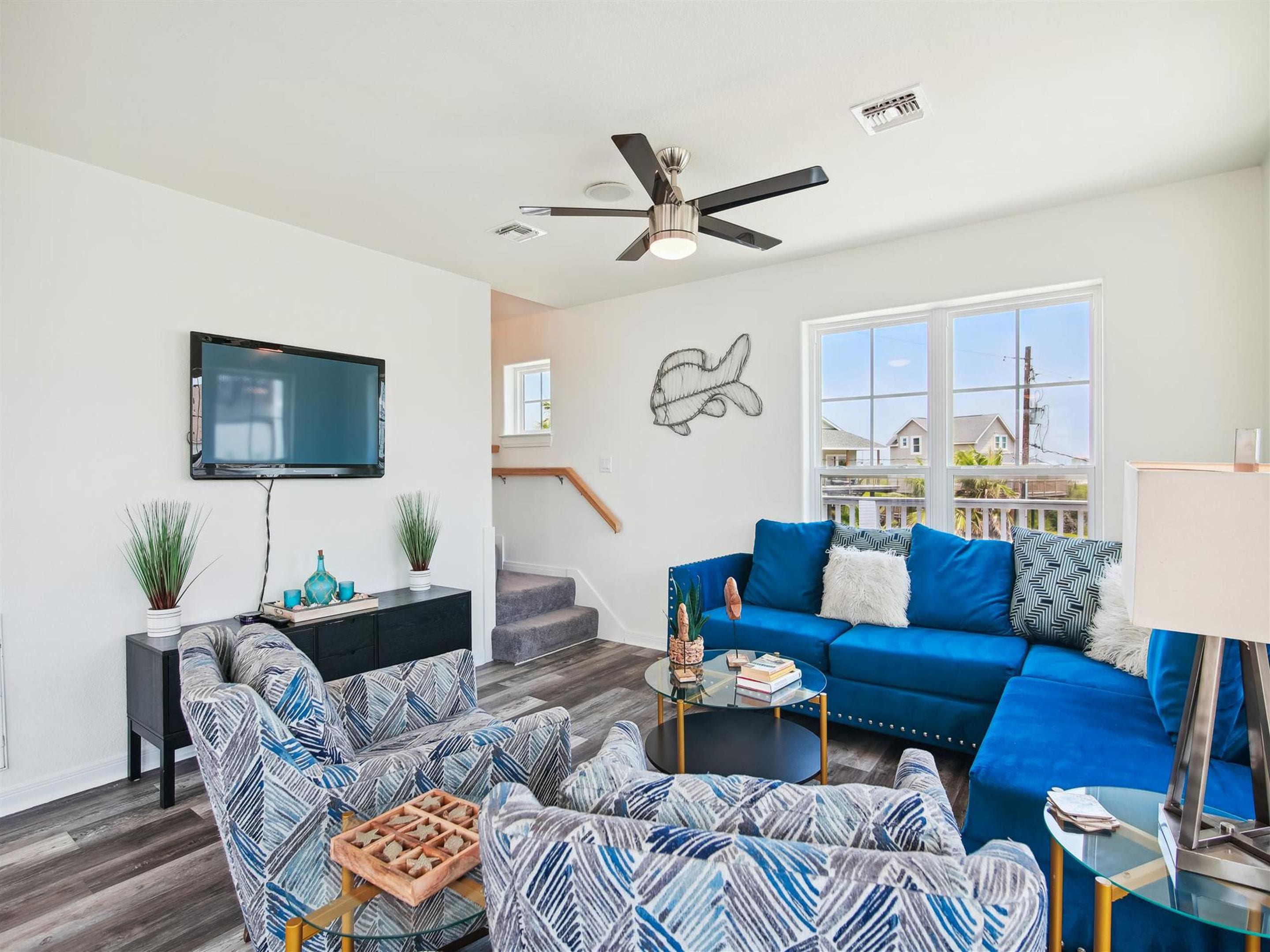 A modern living room with a blue sofa, patterned armchairs, a glass coffee table, and a flat-screen TV mounted on the wall.