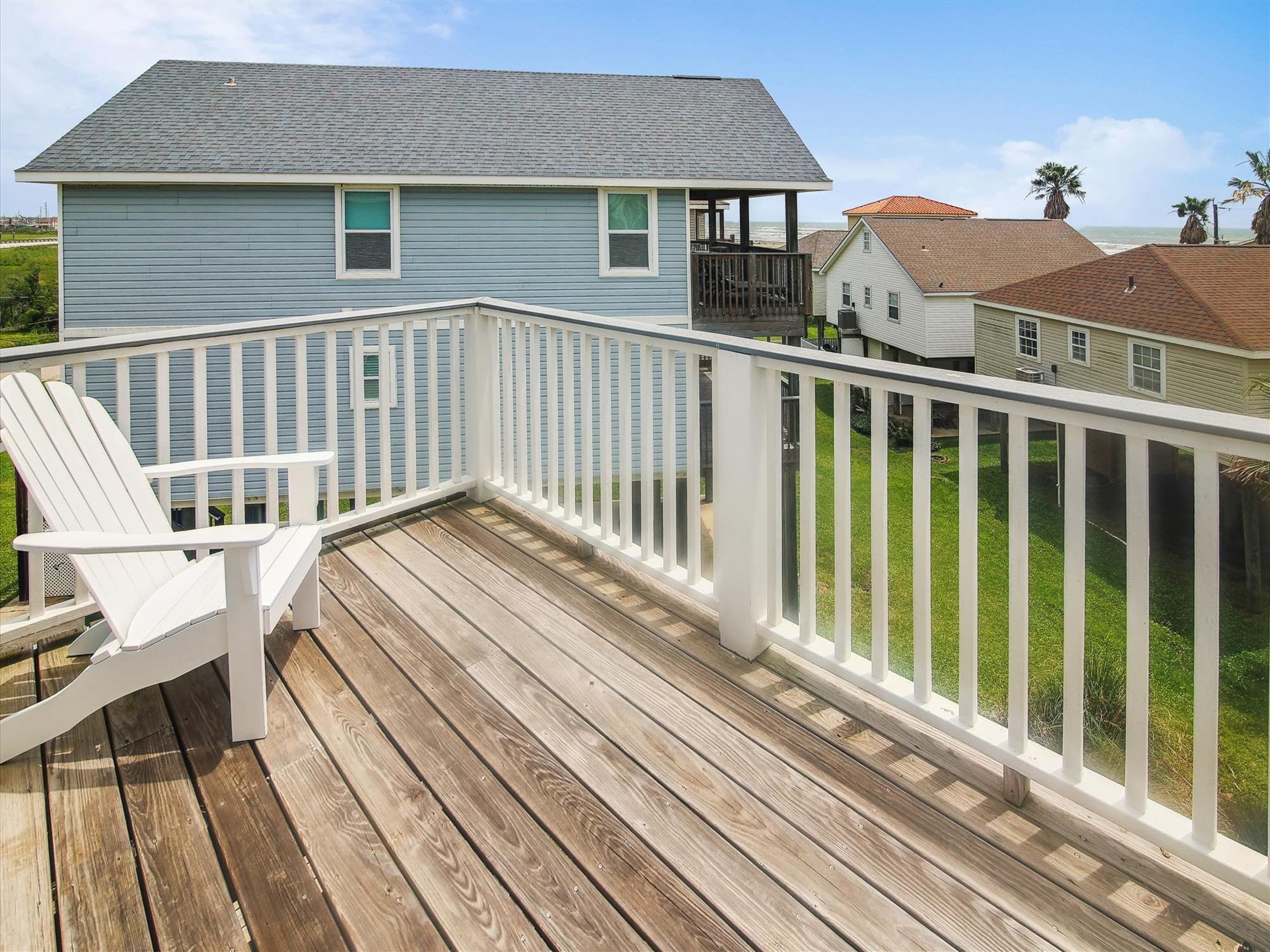 A wooden deck with a white railing, a white Adirondack chair, and a view of a blue house with a grey roof.