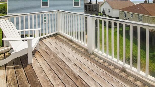A wooden deck with a white railing, a white Adirondack chair, and a view of a blue house with a grey roof.