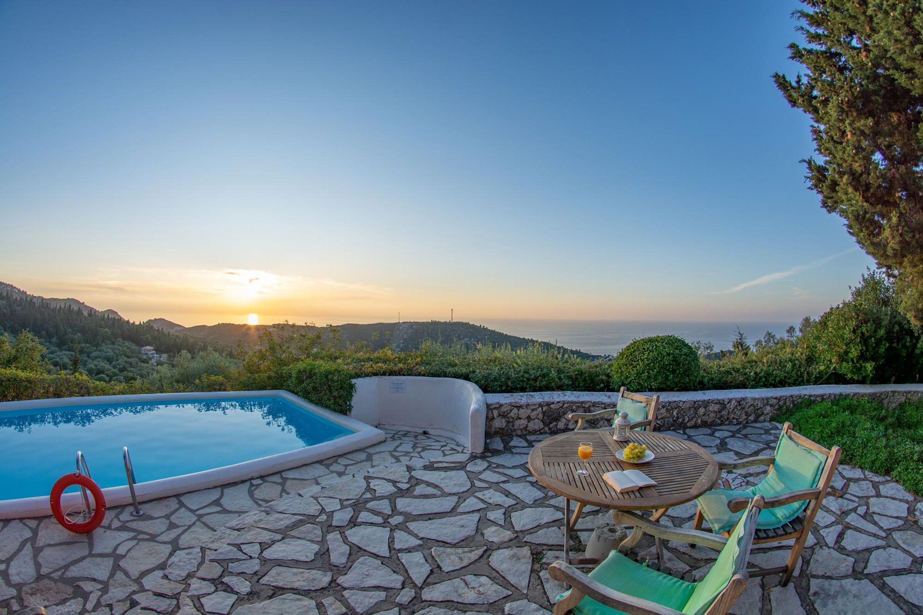 Una piscina de piedra con un flotador salvavidas, rodeada por un patio de piedra con una mesa y sillas, con vistas panorámicas al mar y las colinas durante el atardecer.