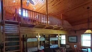 A cozy cabin interior with a loft bedroom, a living area with a sofa and chairs, and a dining area with a table and chairs.