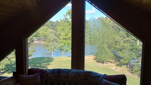 A room with a wooden ceiling, a purple sofa with patterned cushions, and large windows offering a view of a lake and greenery.