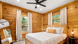 Un dormitorio tipo cabaña de madera con una cama, cortinas, ventilador de techo y una mesita de noche con lámpara.
