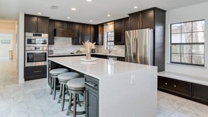 A modern kitchen with dark cabinetry, stainless steel appliances, a large island with bar stools, and a view through a window. - Dolphin View (St. Augustine)