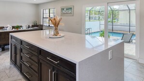 A modern kitchen with dark cabinets, a large island, and a view of a backyard with a pool. - Dolphin View (St. Augustine)