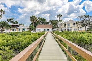 A wooden boardwalk leading to a white building with a red roof, surrounded by lush greenery and palm trees.