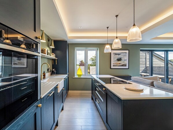 Una cocina moderna con armarios oscuros, una isla central, iluminación colgante y vistas a través de una ventana.