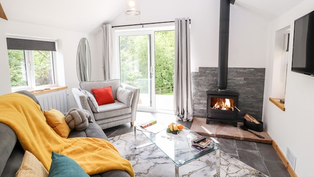 A modern living room with a stone fireplace, a comfortable sofa, a glass coffee table, and a view of greenery outside. - Lands Brow Lodge (Wigton)