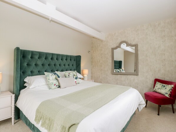 A neatly made bed with a green headboard, white bedding, and a green throw blanket. There is a red chair with a floral pillow, a bedside table with a lamp, and a mirror on the wall.