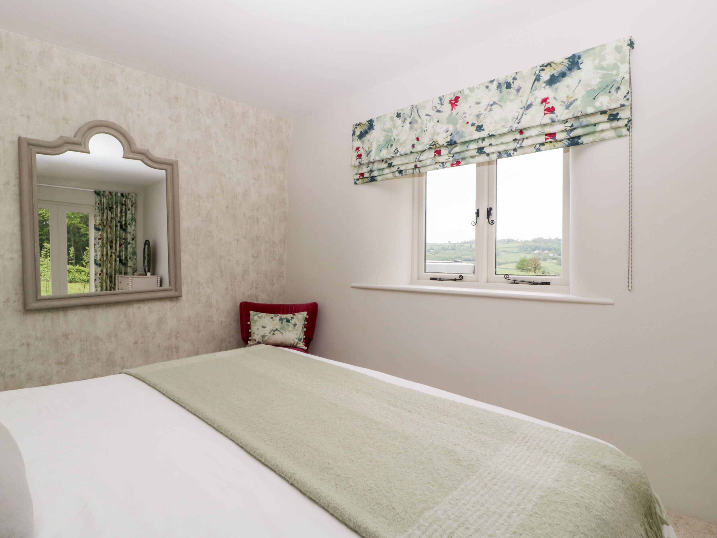 Ein Schlafzimmer mit einem Bett, einem Fenster mit blumigem Sichtschutz, einem Spiegel und einem roten Kissen.