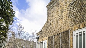 A courtyard with a brick building, white metal chairs and tables, and a small garden area. - The Park Lane Penthouse (London)