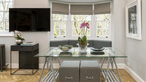 A modern dining area with a glass table, a bench, and a bay window with curtains. - The Park Lane Penthouse (London)