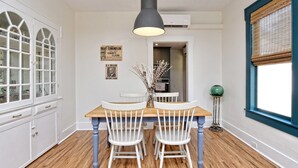 Un comedor con una mesa de madera y sillas blancas, una lámpara colgante y un ventanal con marco azul.