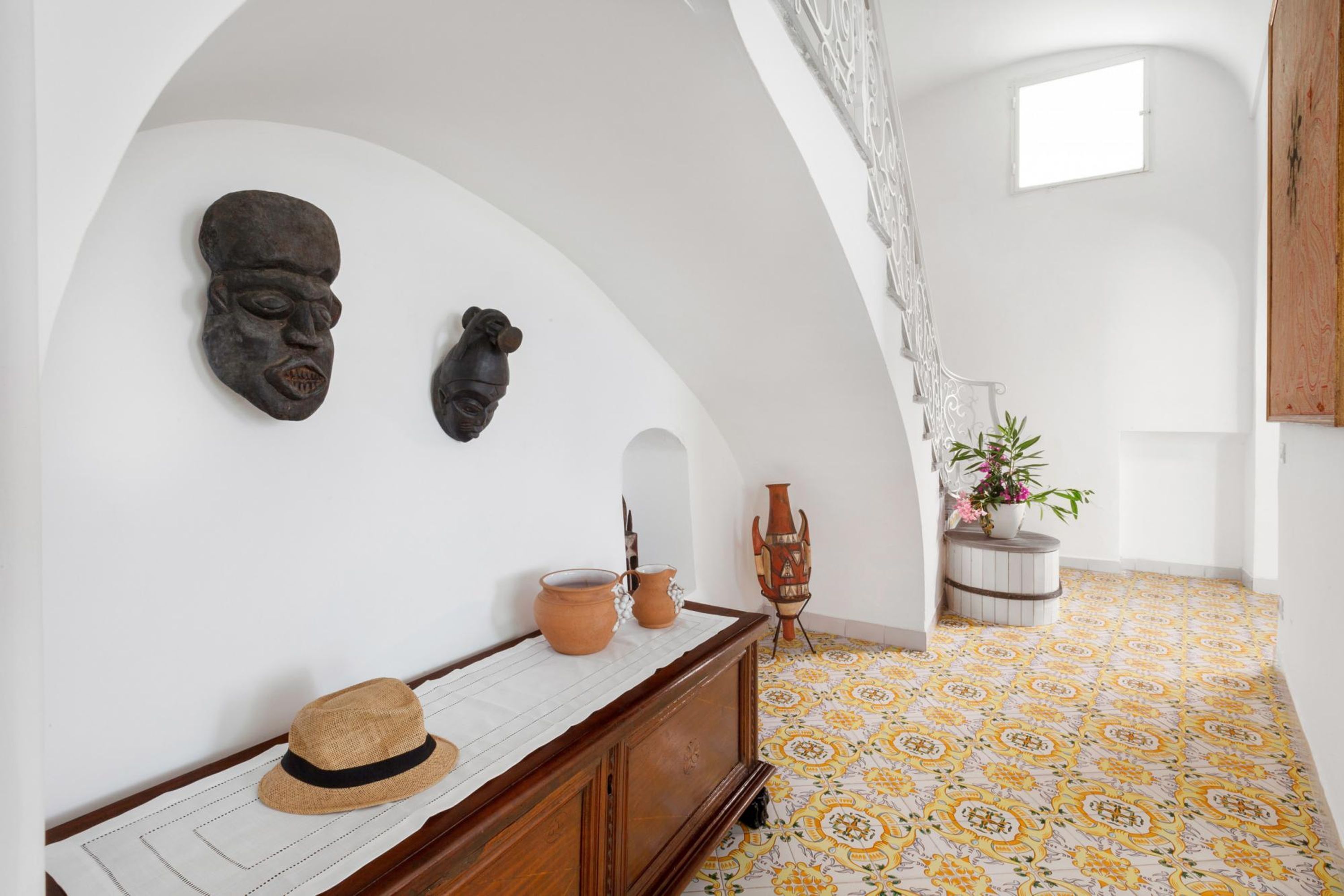 Una habitación con techo blanco curvado, un banco de madera con un sombrero, dos piezas de cerámica y un jarrón con flores.