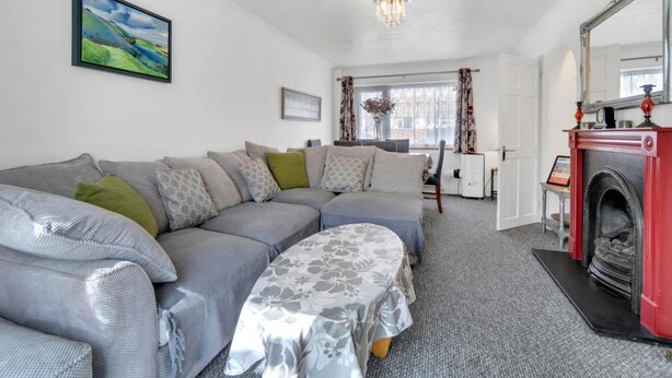 A modern living room with a grey sectional sofa, a small table with a floral tablecloth, a fireplace, and a large window with curtains. - Valley Rise By My Getaways Free Parking (Brighton)