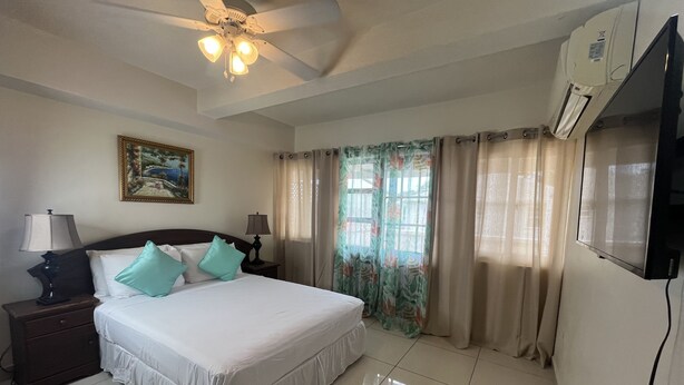 A hotel room with a bed, two bedside tables with lamps, a ceiling fan, a painting on the wall, and a window with curtains. - St. Lawrence Suites - Suite Two (Oistins)