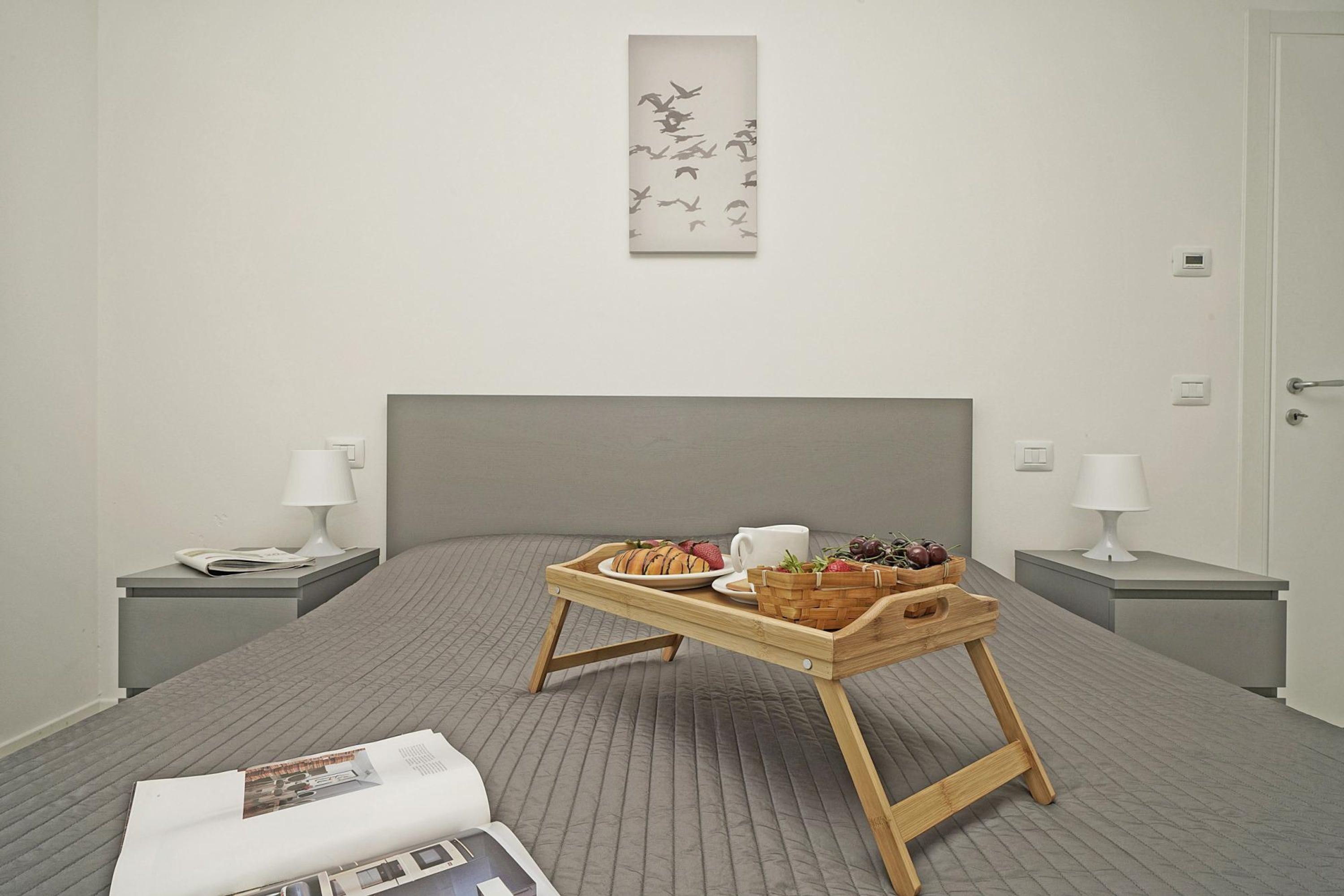 Ein ordentlich eingerichtetes Hotelzimmer mit einem Bett, einem Nachttisch mit Lampe, einer Ablage mit Frühstücksangeboten und einem Wandbild.