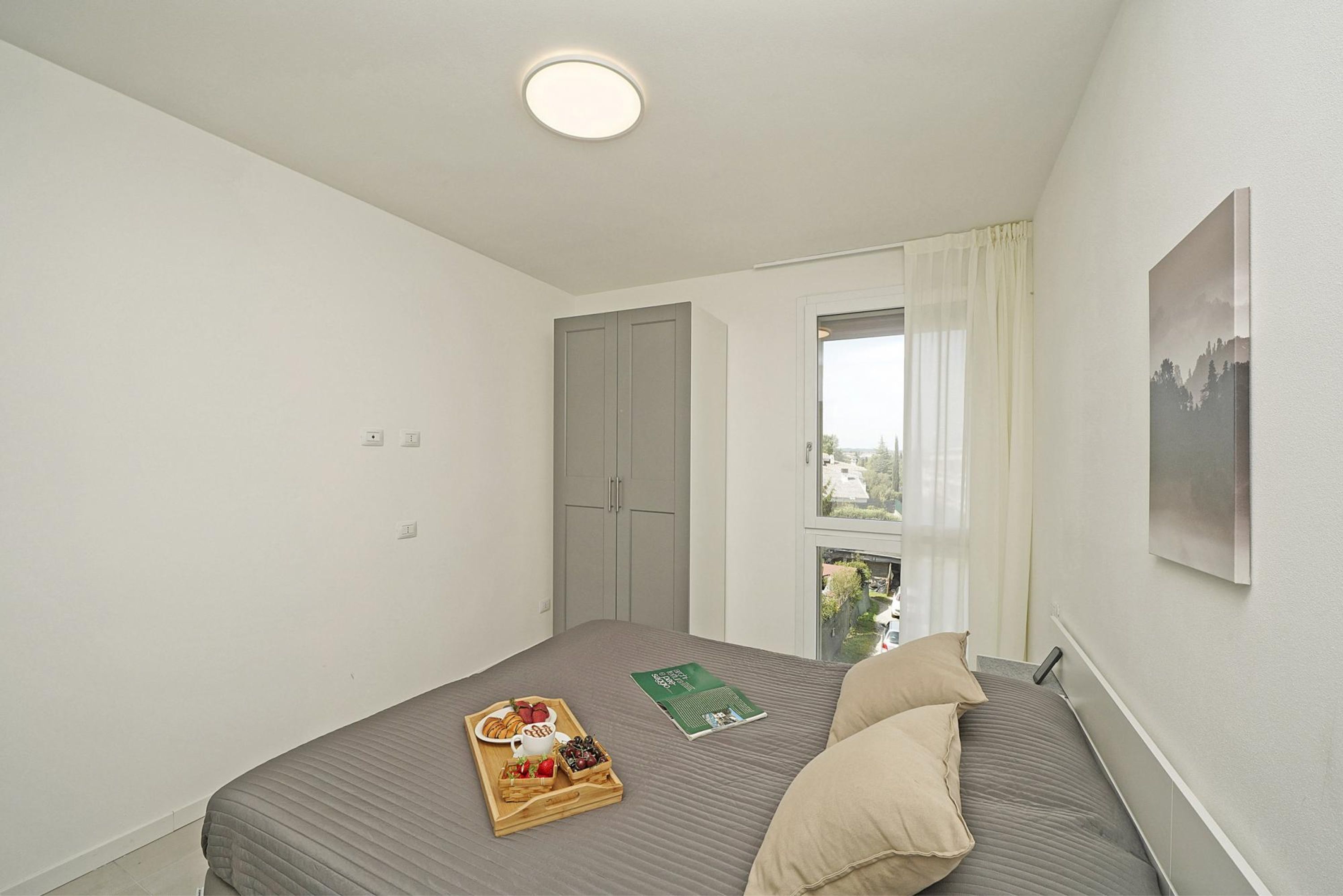 Ein ordentlich eingerichtetes Schlafzimmer mit einem Bett, einem Fenster mit Vorhängen, einem Bild an der Wand und einem Tablett mit Essen auf dem Bett.