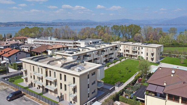 Ein moderner Wohnkomplex mit mehreren Gebäuden, Dachterrassen und Blick auf einen See und Berge im Hintergrund.