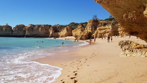 Une plage avec une eau d’un bleu limpide, des falaises rocheuses, et des gens qui se baignent et se promènent sur le sable.