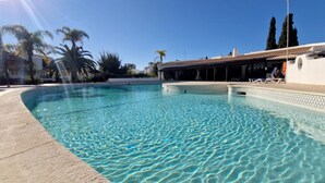 Une piscine aux eaux cristallines, bordée d’une terrasse carrelée, entourée de palmiers et d’un bâtiment doté d’un patio couvert.