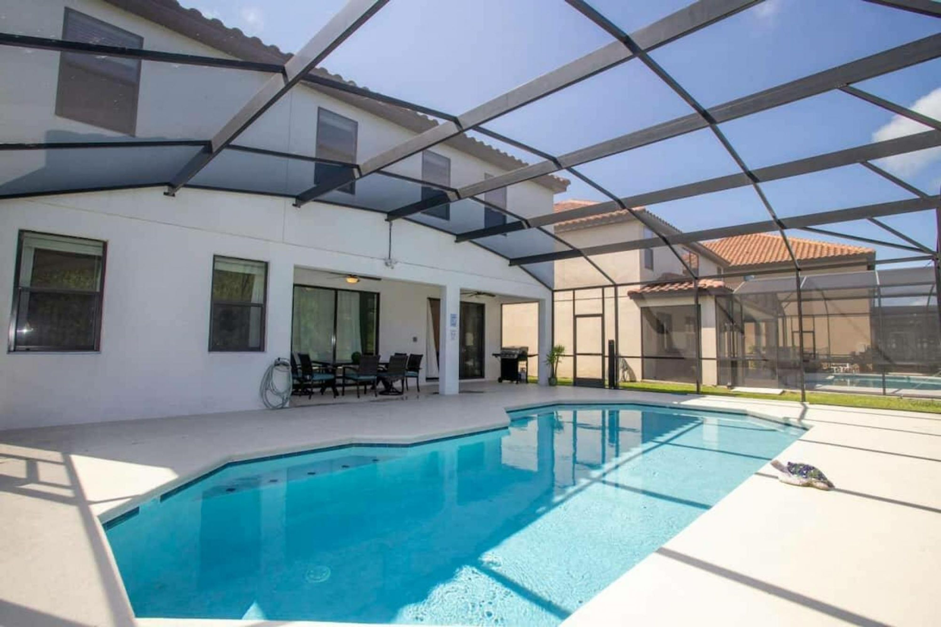 A swimming pool with a glass enclosure, a patio area with chairs, and a view of a building with a tiled roof.