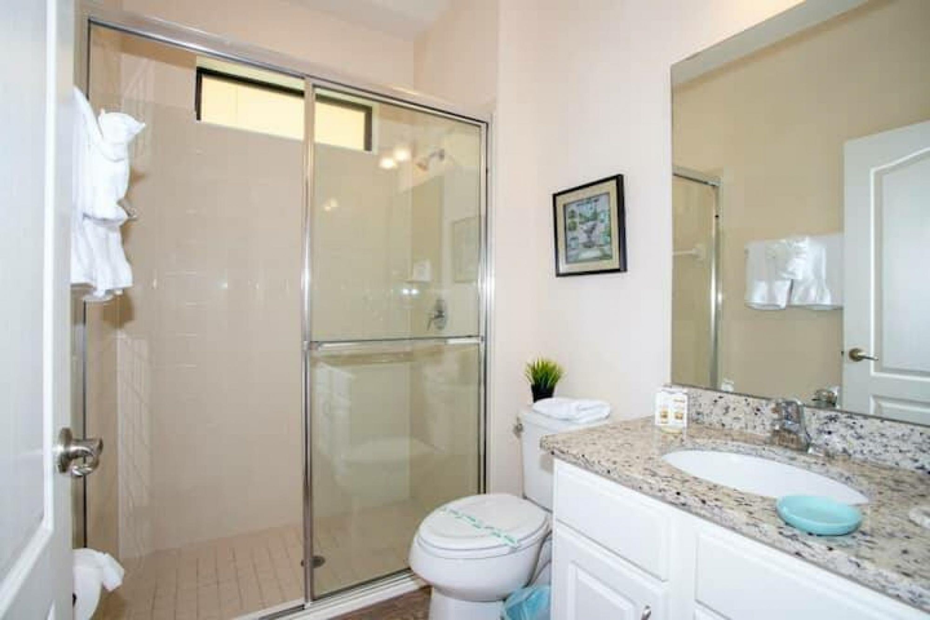 A bathroom with a glass shower enclosure, a white toilet, a sink with a blue soap dish, and a framed picture on the wall.