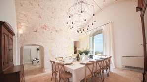A dining area with a large table set for six, surrounded by wooden chairs. A modern chandelier hangs from the ceiling, and a large window provides a view of the outdoors. - TD Tenuta Donna Madia Sea View Pool Over Vineyard (Monopoli)