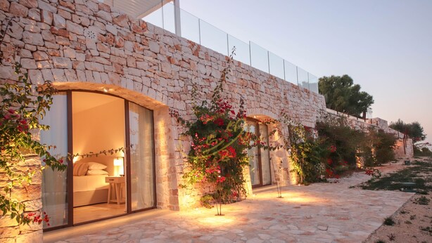A stone building with a glass door entrance, a bed visible inside, and a garden with red flowers. - TD Tenuta Donna Madia Sea View Pool Over Vineyard (Monopoli)
