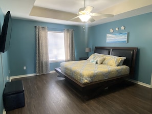 A bedroom with a bed, a ceiling fan, a window with curtains, a nightstand, and a wall-mounted TV.