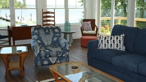 Ein Wohnzimmer mit Blick auf das Wasser, ein blaues Sofa, ein Korbsessel und ein Glastisch.