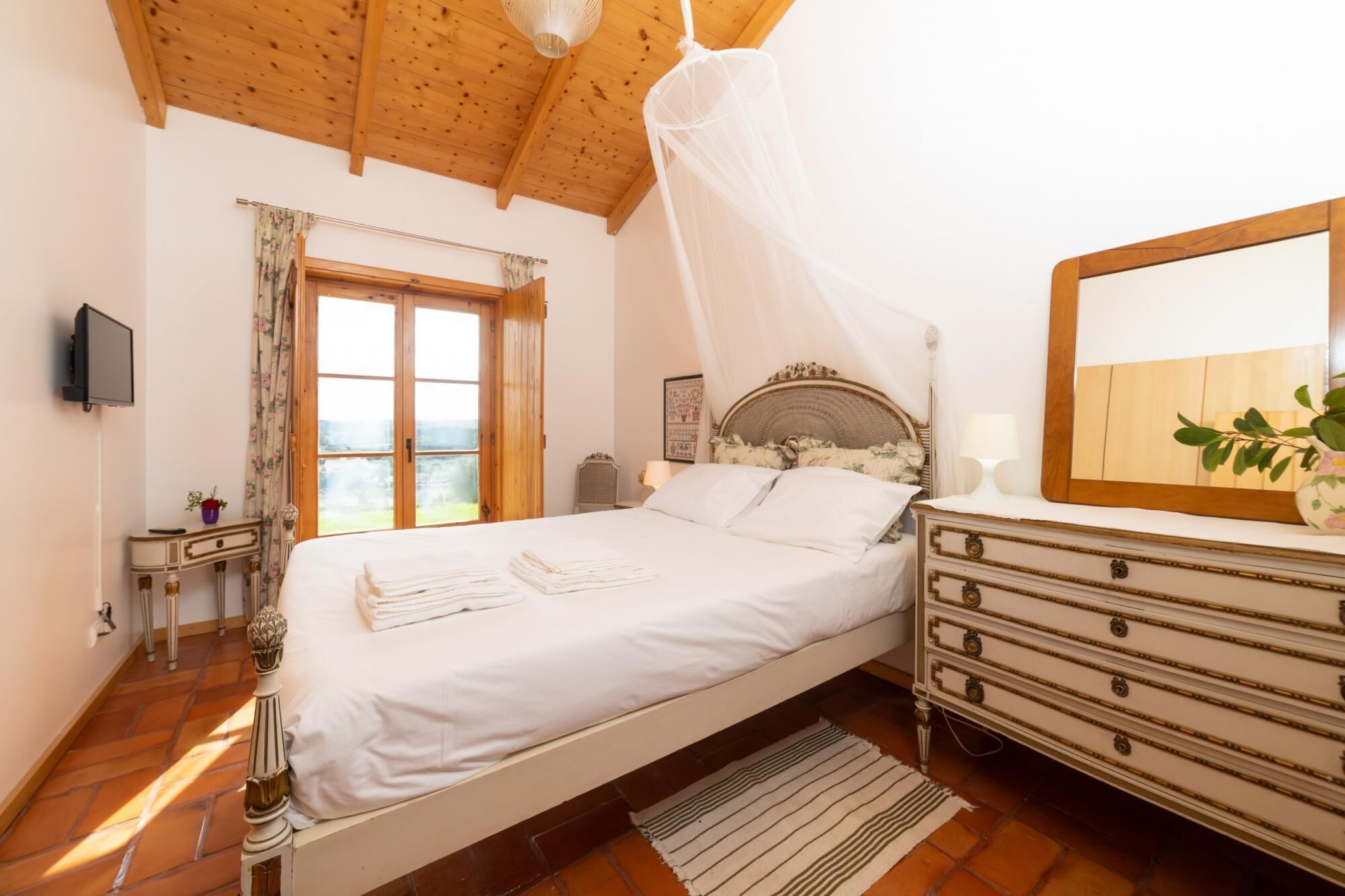 A bedroom with a bed, a dresser, a mirror, a television, and a window with curtains.